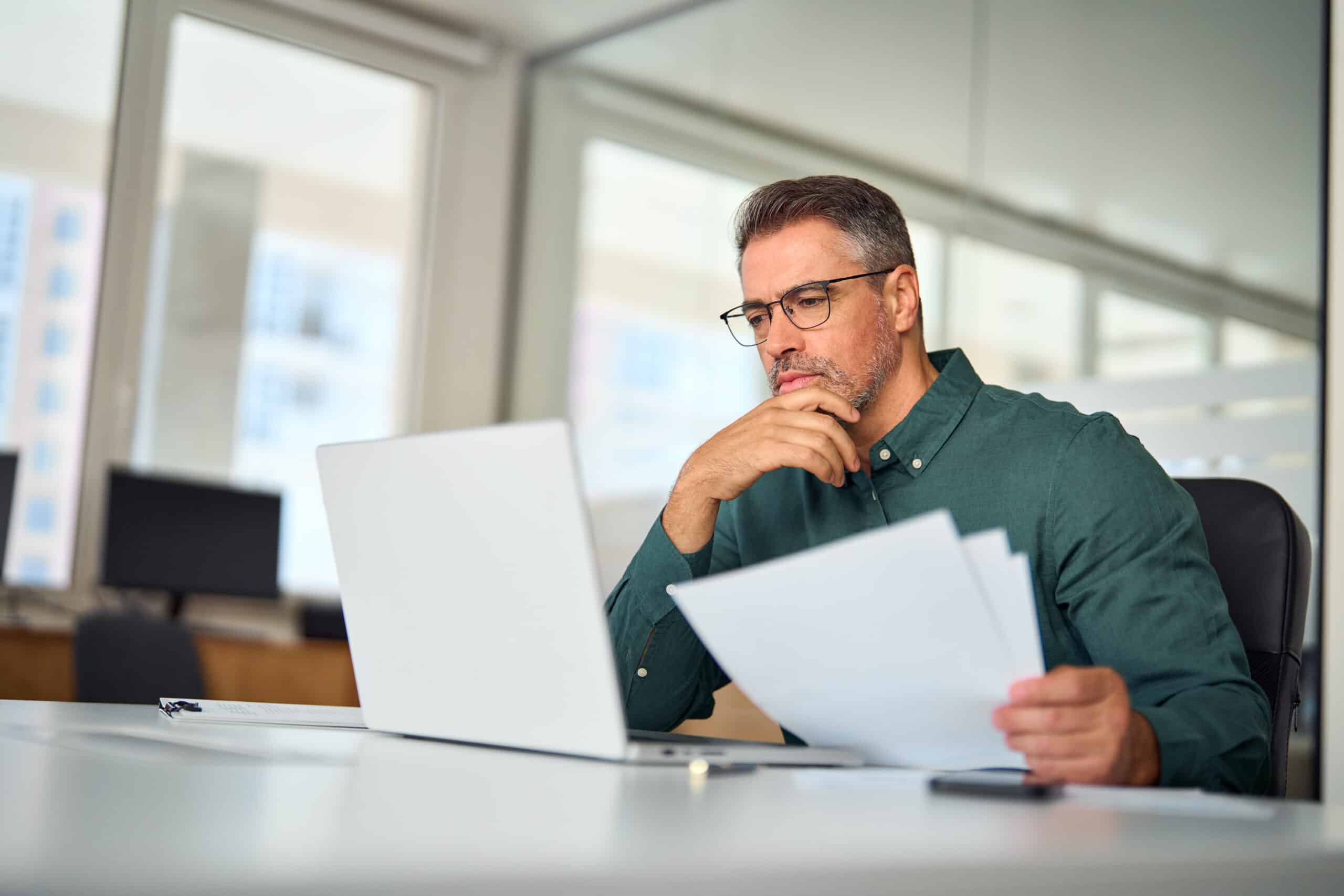 Busy middle aged male hr manager holding documents using laptop looking at reviewing best payroll software options.